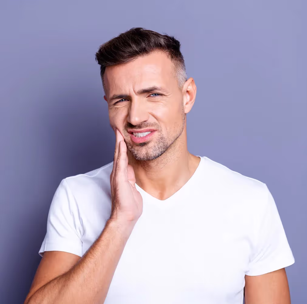 A man with a beard holding his face because he has a toothache. He stands in front of a purple background.